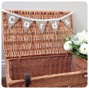 May include: A large, open wicker basket with a hinged lid. A banner reading "cards" hangs above the basket. White flowers and greenery are arranged to the right. The basket has a leather strap and buckle closure.