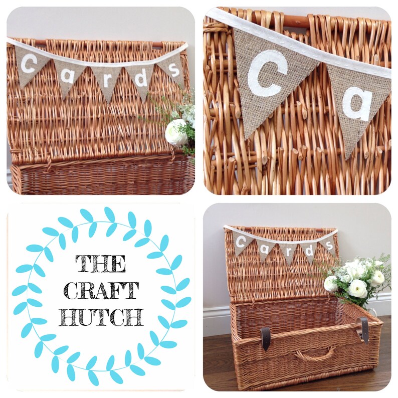 May include: A brown wicker basket with a hinged lid, decorated with a burlap banner that spells "Cards" in white letters. The basket is adorned with white flowers, suggesting a wedding or event theme. The Craft Hutch logo is also visible.