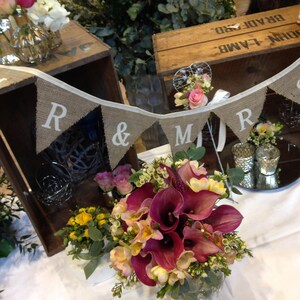 May include: A wedding reception scene with floral arrangements and decorative elements. A burlap banner spells out "R & MRS". The flowers include calla lilies, roses, and other blooms in shades of purple, yellow, and white. Wooden crates and mirrored surfaces are also visible.