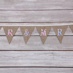 May include: A burlap banner with the words "MR & MRS" in individual pennants. The letters are in a mix of floral and solid colors, including pink and blue. The banner is strung on a cream-colored ribbon.