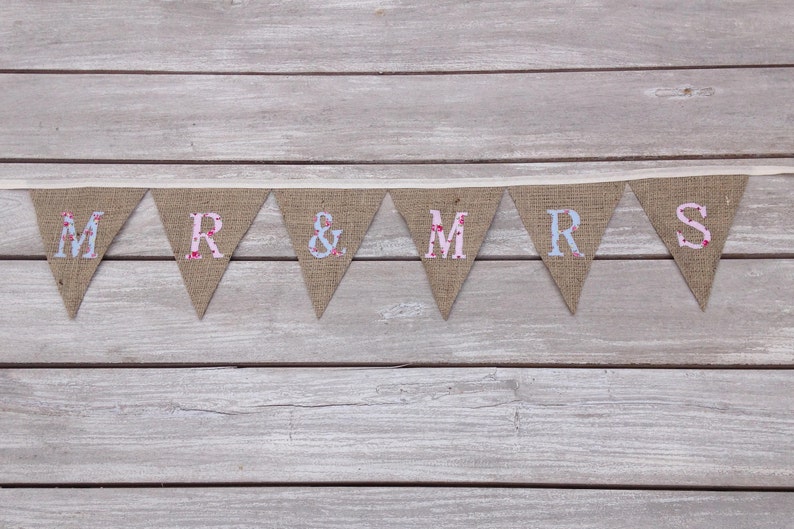 May include: A burlap banner with the words "MR & MRS" in individual pennants. The letters are in a mix of floral and solid colors, including pink and blue. The banner is strung on a cream-colored ribbon.