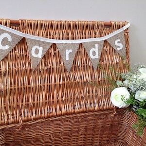 May include: A brown wicker basket with a banner that reads "cards" in white letters. The banner is made of burlap triangles and is strung on a white ribbon. A bouquet of white flowers is to the right of the basket.