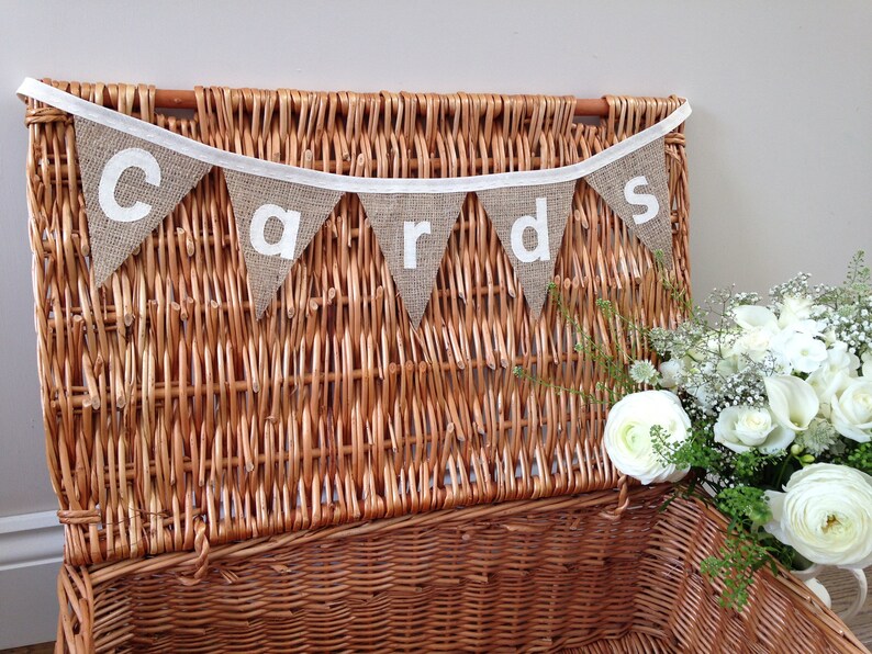 May include: A brown wicker basket with a banner that reads "cards" in white letters. The banner is made of burlap triangles and is strung on a white ribbon. A bouquet of white flowers is to the right of the basket.