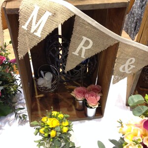 May include: A rustic wooden crate with a burlap banner that reads "MR &" is decorated with flowers and decorative items. The crate is filled with pink roses in small metal containers, a wire basket, and a decorative sphere. A small bouquet of yellow flowers sits in front.