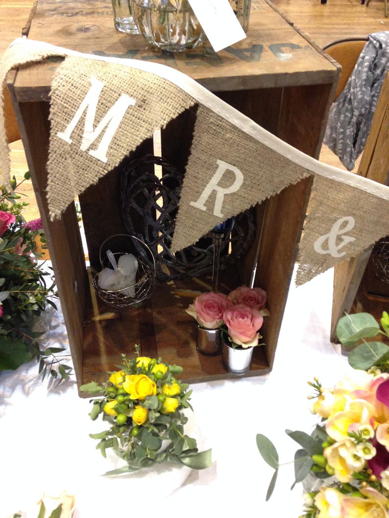 May include: A rustic wooden crate with a burlap banner that reads "MR &" is decorated with flowers and decorative items. The crate is filled with pink roses in small metal containers, a wire basket, and a decorative sphere. A small bouquet of yellow flowers sits in front.