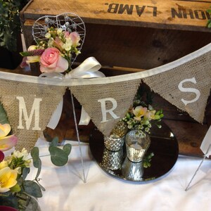 May include: A burlap banner with the letters "MRS" in white, strung across a wooden crate. The banner is decorated with flowers and a heart-shaped wire decoration. A mirrored surface reflects candles and floral arrangements.