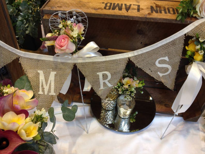 May include: A burlap banner with the letters "MRS" in white, strung across a wooden crate. The banner is decorated with flowers and a heart-shaped wire decoration. A mirrored surface reflects candles and floral arrangements.