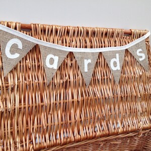 May include: A rustic burlap banner with the word "cards" in white letters hangs above a woven wicker basket. The banner is made of triangular flags with white trim and is suspended from a white ribbon. The basket is a warm brown color.