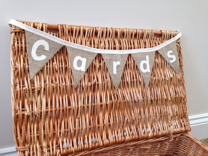 May include: A rustic burlap banner with the word "cards" in white letters hangs above a woven wicker basket. The banner is made of triangular flags with white trim and is suspended from a white ribbon. The basket is a warm brown color.