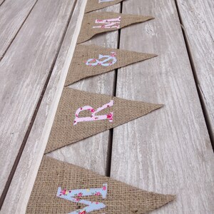 May include: A burlap banner with pennant flags spelling out "MRS &" in floral print letters. The banner is strung on a white ribbon and rests on a weathered wooden surface. The letters are in pink and blue floral patterns.