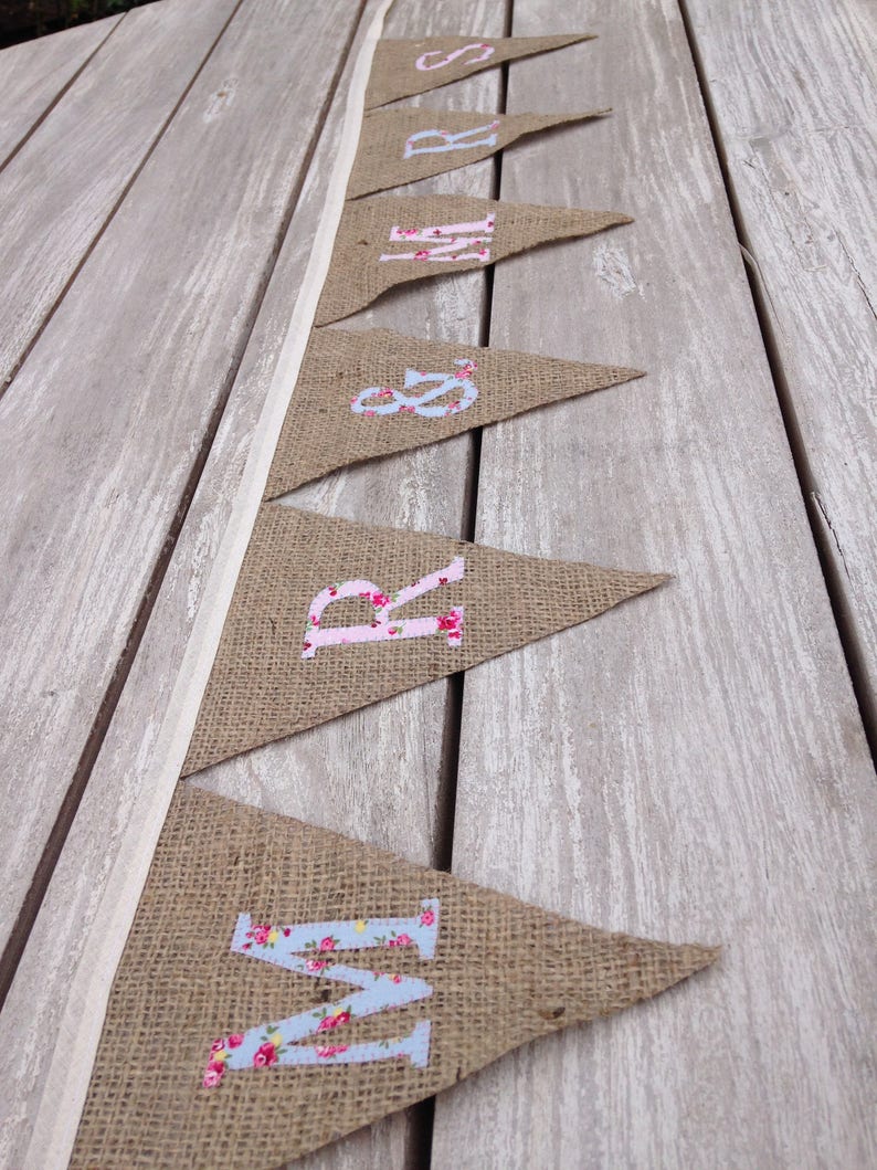 May include: A burlap banner with pennant flags spelling out "MRS &" in floral print letters. The banner is strung on a white ribbon and rests on a weathered wooden surface. The letters are in pink and blue floral patterns.