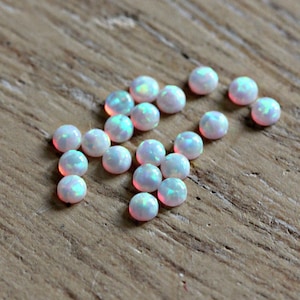 May include: A group of 18 small, round, white opal cabochons with iridescent rainbow colors. The opals are scattered on a wooden surface.