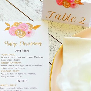 May include: A white menu card with gold lettering for a christening celebration. The menu features appetizers and entrees, including Verde Salad, Grilled Flatbread, Avocado Toast, and Huevos 'Bulla'.