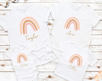 May include: Four white t-shirts with a pink and brown rainbow design and a name printed below each rainbow. The names are Taylor, Ava, Riley, and Ella.