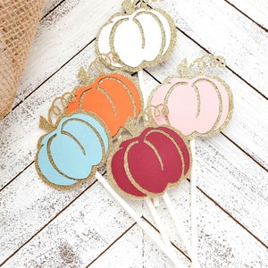 May include: Six glitter pumpkin cupcake toppers in various colors, including white, orange, pink, blue, and red. The toppers are attached to white sticks.