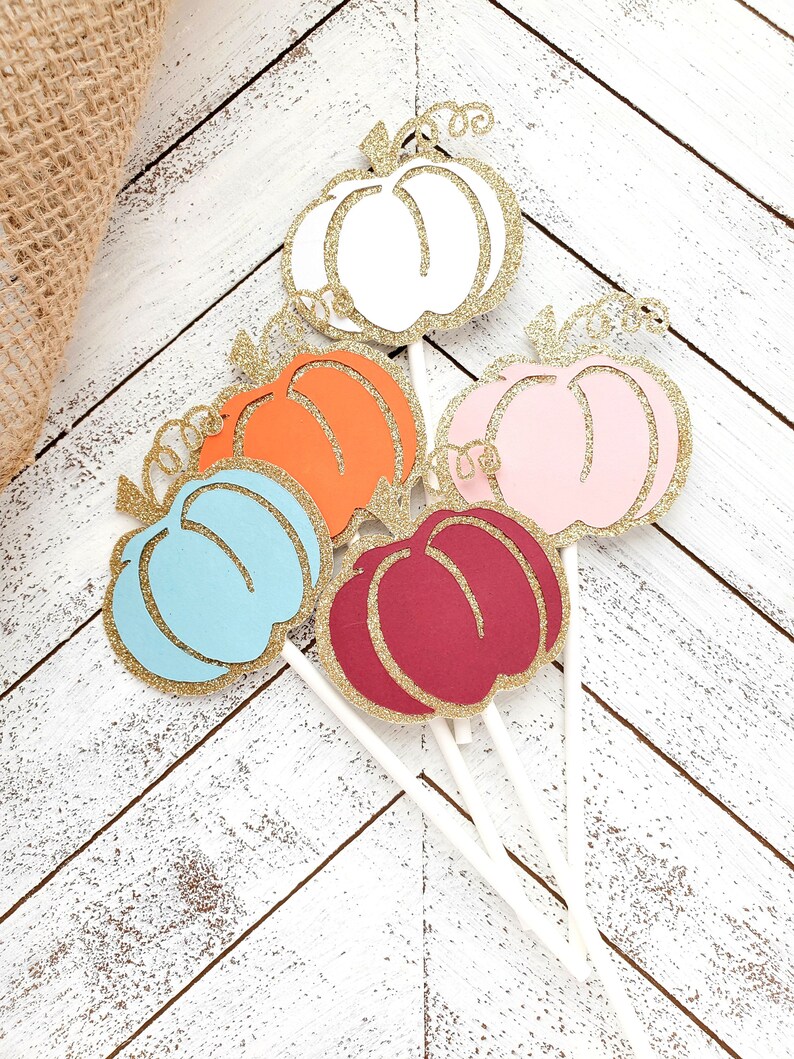 May include: Six glitter pumpkin cupcake toppers in various colors, including white, orange, pink, blue, and red. The toppers are attached to white sticks.