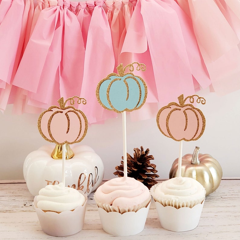 May include: Three cupcakes decorated with gold glitter pumpkin toppers. The cupcakes are topped with white, pink, and light blue frosting. The pumpkins are blue, pink, and light blue. The cupcakes are sitting on a white surface with a pink and white tissue paper backdrop.