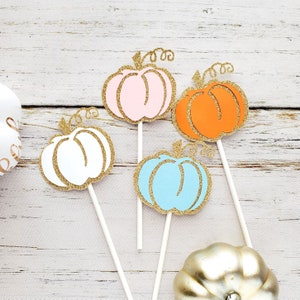 May include: Four pumpkin-shaped cake toppers with gold glitter accents. The toppers are in pink, white, blue, and orange. Each topper has a stick for inserting into a cake.