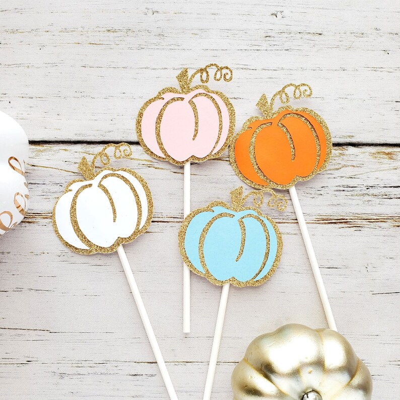 May include: Four pumpkin-shaped cake toppers with gold glitter accents. The toppers are in pink, white, blue, and orange. Each topper has a stick for inserting into a cake.