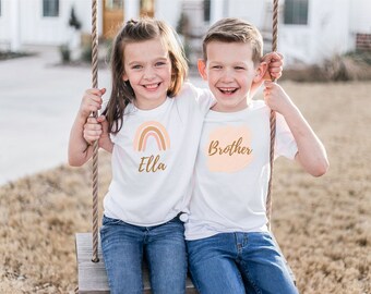 May include: Two children on a swing, wearing white t-shirts with a rainbow and the name "Ella" and the word "Brother" printed on them. The children are smiling and looking at the camera.