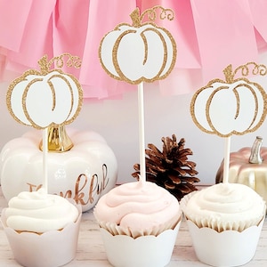 May include: Three white cupcakes with pink and white frosting topped with gold glitter pumpkin cupcake toppers. The cupcakes are on a wooden surface with a white pumpkin and a pine cone.