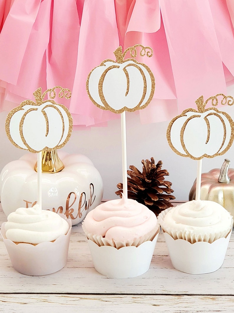 May include: Three white cupcakes with pink and white frosting topped with gold glitter pumpkin cupcake toppers. The cupcakes are on a wooden surface with a white pumpkin and a pine cone.