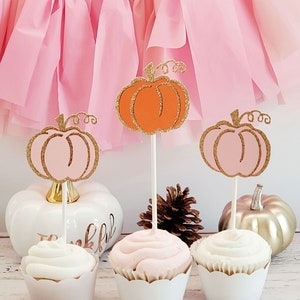 May include: Three cupcakes decorated with pink and orange glitter pumpkin toppers. The cupcakes are frosted with white and pink frosting. The cupcakes are on a white surface.