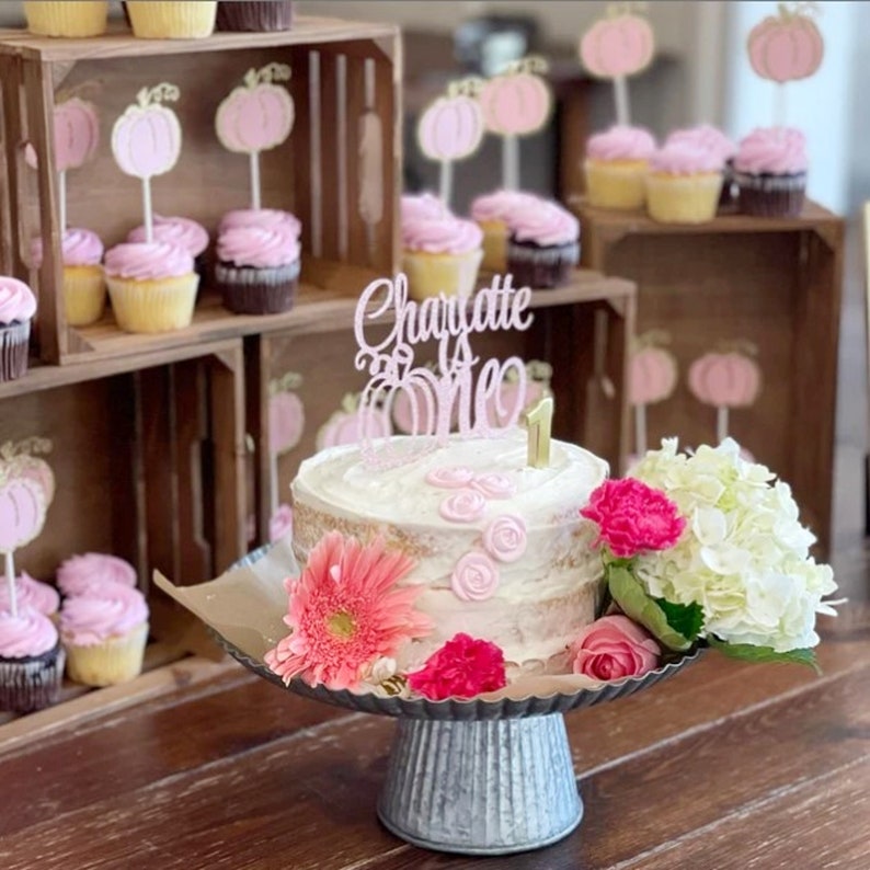 May include: A white cake with pink frosting and a pink and gold "Charlotte is One" cake topper. The cake is decorated with pink roses and is on a silver cake stand. The cake is surrounded by pink and white flowers.