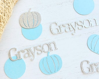 May include: Scattered on a white surface are blue and silver pumpkin-shaped paper cutouts. The name "Grayson" is spelled out in silver glitter letters twice.