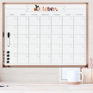 Puede incluir: Un calendario de borrado en seco blanco con un marco negro y un borde de oro rosa. El calendario est&aacute; etiquetado como "Octubre" con un gr&aacute;fico de bruja y calabaza. El calendario tiene una cuadr&iacute;cula de cuadrados para cada d&iacute;a del mes, con los d&iacute;as de la semana etiquetados en la parte superior.
