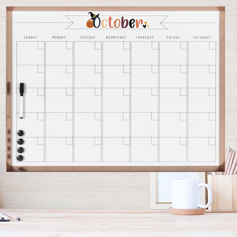 Puede incluir: Un calendario de borrado en seco blanco con un marco negro y un borde de oro rosa. El calendario est&aacute; etiquetado como "Octubre" con un gr&aacute;fico de bruja y calabaza. El calendario tiene una cuadr&iacute;cula de cuadrados para cada d&iacute;a del mes, con los d&iacute;as de la semana etiquetados en la parte superior.