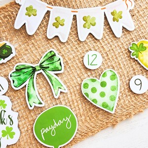 May include: Green and white St. Patrick's Day themed decorations on a burlap background. The decorations include a banner, a bow, a heart, a pot of gold, and numbers 9 and 12. The banner has shamrocks and the text "Payday".