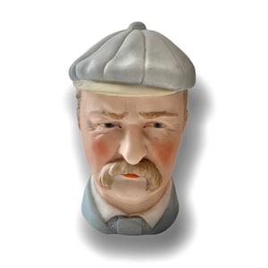 May include: A porcelain figurine of a man with a grey cap, moustache, and beard. He is wearing a grey shirt and has a serious expression.