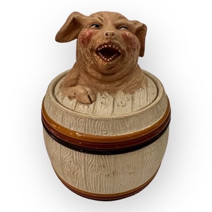 Puede incluir: Una alcancía de cerámica con forma de cerdo en un barril. El cerdo es marrón y tiene un hocico blanco y una amplia sonrisa. El barril es blanco con rayas marrones.