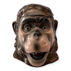 May include: A vintage ceramic monkey cookie jar. The jar is brown with a light pink face and open mouth. The monkey has large, expressive eyes and a detailed fur texture. The lid sits on top of the monkey's head.