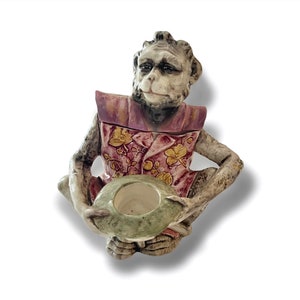 May include: A ceramic monkey figurine wearing a pink and purple patterned shirt, holding a green bowl with a white interior. The monkey has a realistic face with brown eyes and a closed mouth.