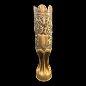 May include: A vintage brass vase with ornate detailing. The vase features embossed text and a shield design, with the year 1918 visible. The vase has a flared base and a crown-like top, showcasing a detailed, aged aesthetic.