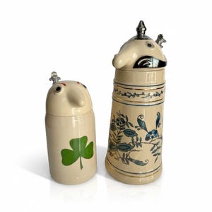 May include: Two cream-colored ceramic steins with lids. The shorter stein features a green shamrock design. The taller stein is decorated with blue and green floral patterns, gold trim, and a metallic lid topper shaped like a bird figure.