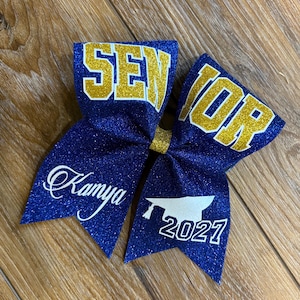 May include: A blue glitter cheer bow with gold lettering that says "SENIOR" and "2027". The bow also has the name "Kamya" in white script. A graduation-themed accessory.