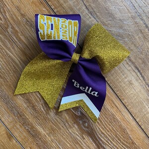 May include: A large hair bow featuring purple and gold glitter fabric. The top loop displays the word SENIOR in gold glitter lettering. One of the bottom tails features the name Bella in white script, accented by a white and gold chevron stripe.