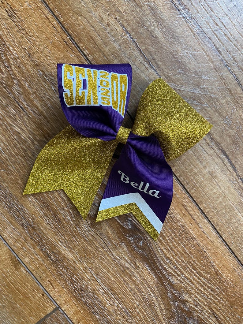 May include: A large hair bow featuring purple and gold glitter fabric. The top loop displays the word SENIOR in gold glitter lettering. One of the bottom tails features the name Bella in white script, accented by a white and gold chevron stripe.
