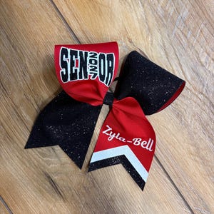 May include: A large hair bow featuring red and black glitter fabric. The top loop displays the word SENIOR in white block letters with black outlines. One tail features the name Zyla-Bell in white script above a white and black chevron pattern.