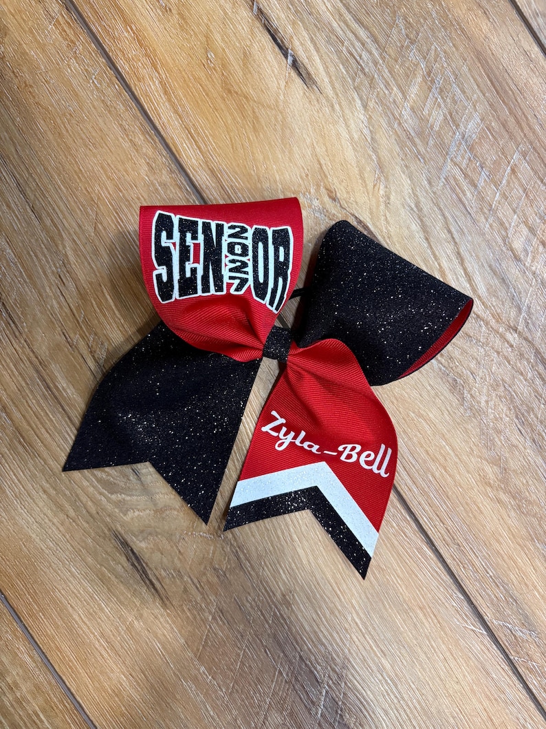 May include: A large hair bow featuring red and black glitter fabric. The top loop displays the word SENIOR in white block letters with black outlines. One tail features the name Zyla-Bell in white script above a white and black chevron pattern.