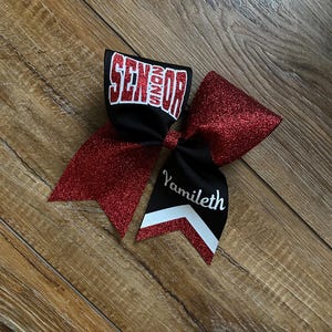 May include: A hair bow featuring red glitter fabric and black ribbon. The top loop displays the word SENIOR in white block letters with a red outline. The bottom tail features the name Yamileth in white script above a white and red chevron pattern.