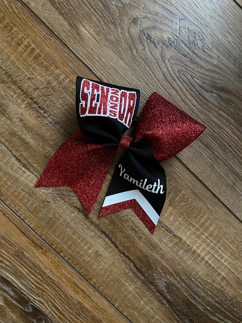 May include: A hair bow featuring red glitter fabric and black ribbon. The top loop displays the word SENIOR in white block letters with a red outline. The bottom tail features the name Yamileth in white script above a white and red chevron pattern.