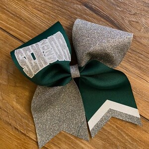 May include: A hair bow constructed from dark green grosgrain ribbon and silver glitter fabric. The left loop features the word SENIOR in silver glitter lettering. The right tail displays a chevron pattern with white and silver glitter stripes.