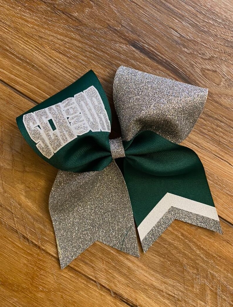 May include: A hair bow constructed from dark green grosgrain ribbon and silver glitter fabric. The left loop features the word SENIOR in silver glitter lettering. The right tail displays a chevron pattern with white and silver glitter stripes.