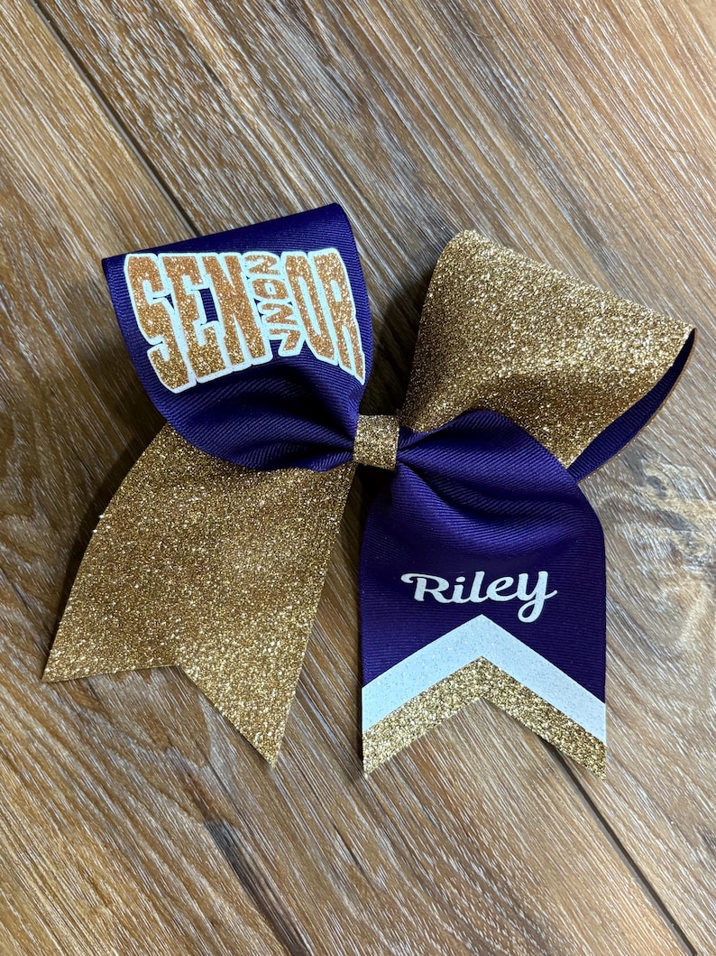May include: A purple and gold glitter hair bow featuring the word SENIOR in gold lettering on the left loop and the name Riley in white script above a white and gold chevron pattern on the right tail, constructed from grosgrain ribbon.