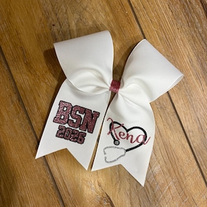 May include: White cheer bow with a pink glitter accent. One side features "BSN 2026" in pink glitter, the other side has a heart with a stethoscope and the name "Kena" in pink.