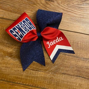 May include: A cheer bow featuring a combination of red fabric and dark blue glitter fabric. The left side displays the word SENIOR in dark blue glitter lettering, while the right side features the name Jaeda in white script with a white chevron accent.